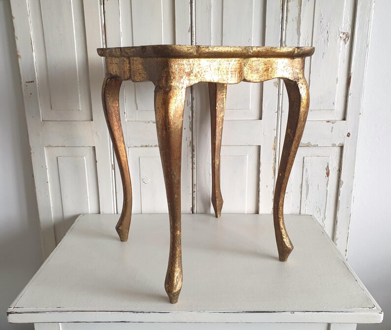 May include: A small, antique gold-colored table with a scalloped edge. The table has four curved legs. The table is approximately 20 inches in diameter and 24 inches tall. The table is made of wood and has a distressed finish.