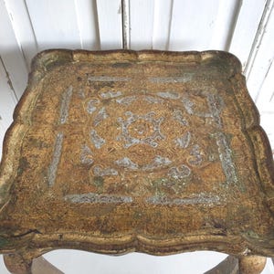 May include: An antique, ornate serving tray with a gold and silver finish. The tray has a scalloped edge and a detailed, symmetrical design in the center. The tray's surface has a distressed appearance, adding to its vintage charm. The tray is supported by four legs.