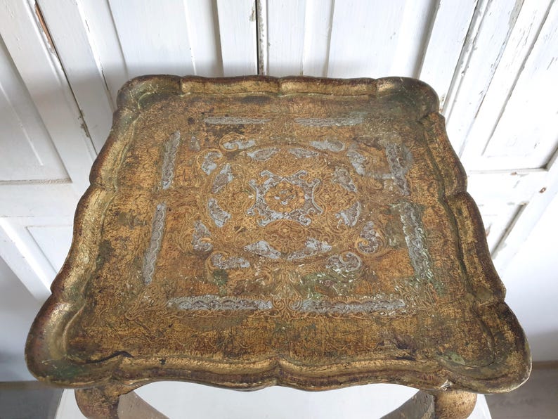 May include: An antique, ornate serving tray with a gold and silver finish. The tray has a scalloped edge and a detailed, symmetrical design in the center. The tray's surface has a distressed appearance, adding to its vintage charm. The tray is supported by four legs.