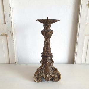 May include: Ornate bronze candlestick holder with a detailed floral and scroll design. The candle holder has a wide, decorative base and a slender, textured stem. The top features a circular dish to hold a candle. A vintage home decor item.
