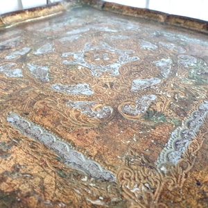 May include: A vintage, square tray with an ornate, embossed design. The tray features a distressed, aged appearance with a gold and brown color palette, accented by areas of faded white and green. The decorative pattern includes floral and scroll motifs.