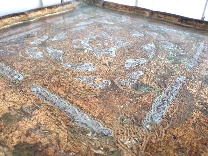 May include: A vintage, square tray with an ornate, embossed design. The tray features a distressed, aged appearance with a gold and brown color palette, accented by areas of faded white and green. The decorative pattern includes floral and scroll motifs.