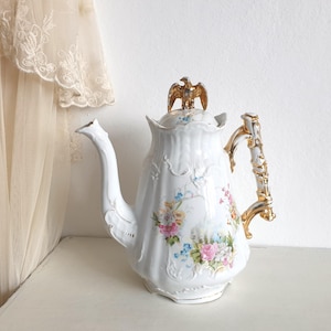 May include: A white porcelain coffee pot with a gold eagle finial and handle. The pot features a floral design in pink, blue, and yellow. The handle is gold and white, and the spout is curved. The pot is on a white surface.