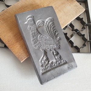 Könnte beinhalten: Eine antike Metallform zum Backen eines Kuchen in Form eines Hahns. Die Form ist aus dunkelgrauem Metall und hat ein detailliertes Design eines Hahns mit einem hochgestellten Schwanz und einem Kamm.