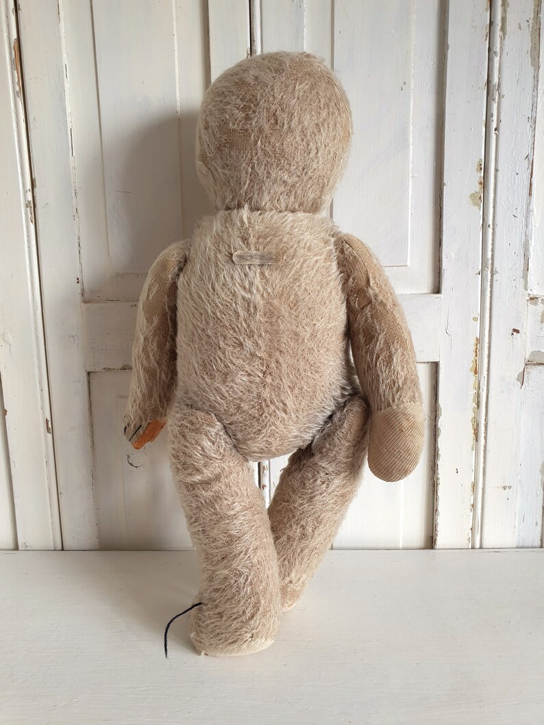 May include: A vintage, beige, mohair teddy bear with a button nose and stitched eyes. The bear is standing with its legs slightly apart and its arms at its sides. The bear has a tag on its back.