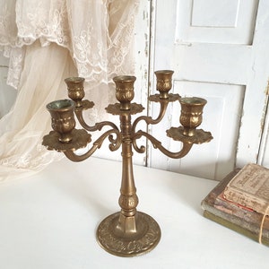 May include: A vintage brass candelabra with five arms, each holding a candle cup. The candelabra has a detailed floral design on the base and arms. The overall height is approximately 12 inches.