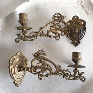 May include: Two antique brass wall sconces. Each sconce features a candle holder, ornate scrollwork, and a decorative backplate. The sconces are made of brass and have a warm, aged patina. They are designed to be mounted on a wall.