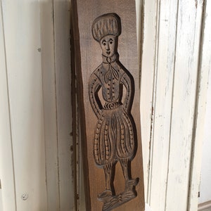 Puede incluir: Un molde de madera rectangular vintage con un relieve tallado de una figura con vestimenta hist&oacute;rica. La figura lleva un sombrero, una chaqueta abotonada y pantalones abombados. La madera tiene un tono marr&oacute;n c&aacute;lido y los detalles del tallado est&aacute;n bien definidos.
