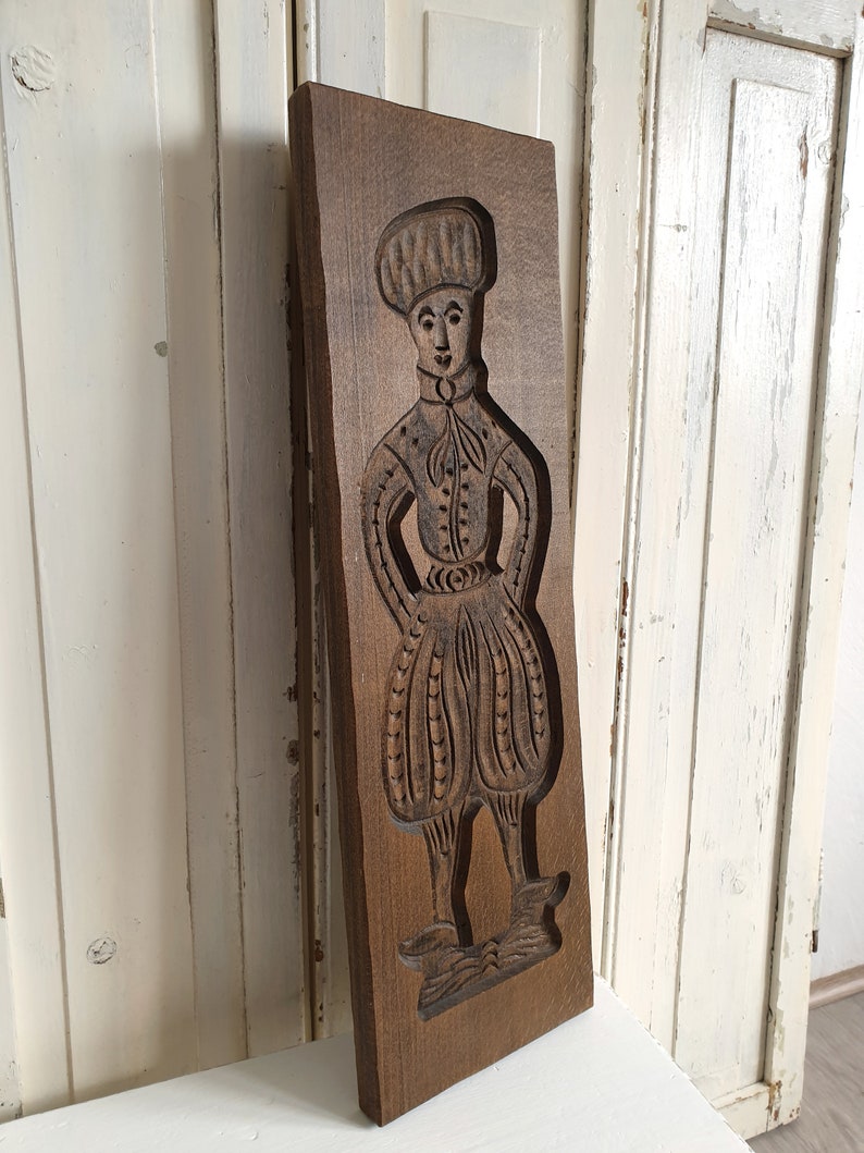 Puede incluir: Un molde de madera rectangular vintage con un relieve tallado de una figura con vestimenta hist&oacute;rica. La figura lleva un sombrero, una chaqueta abotonada y pantalones abombados. La madera tiene un tono marr&oacute;n c&aacute;lido y los detalles del tallado est&aacute;n bien definidos.