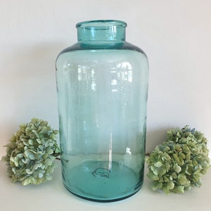May include: A large, clear, teal-colored glass jar with a wide mouth. The jar is sitting on a white surface with two green hydrangea flower clusters on either side.