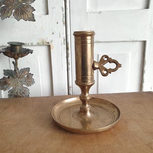May include: A vintage brass candlestick holder with a round base and a cylindrical candle cup. The holder features an ornate handle and is approximately 6 inches tall. The brass has a warm, aged patina, suggesting its antique nature.