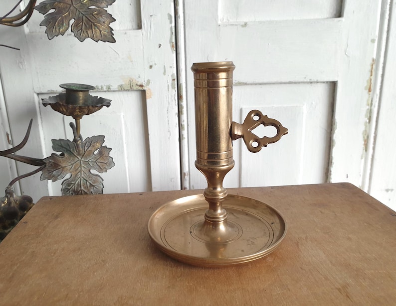 May include: A vintage brass candlestick holder with a round base and a cylindrical candle cup. The holder features an ornate handle and is approximately 6 inches tall. The brass has a warm, aged patina, suggesting its antique nature.
