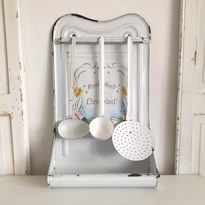 Puede incluir: Un estante de utensilios de cocina de esmalte blanco con diseño floral y el texto "Reinheit Ehre Kleid!" colgado en una pared blanca. El estante tiene tres ganchos y sostiene una cuchara de servir blanca, una cuchara de servir blanca y una cuchara ranurada blanca.