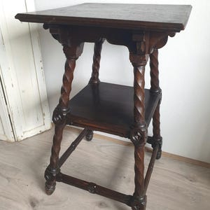 May include: Dark brown antique side table with a square top and a lower shelf. The table features intricately carved, twisted legs and decorative accents. The table's dimensions are approximately 24 inches tall, 18 inches wide, and 18 inches deep.