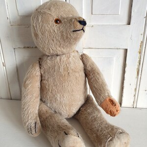 May include: A vintage beige teddy bear with black eyes and a stitched nose. The bear is sitting with its legs crossed and has a worn patch on one arm.