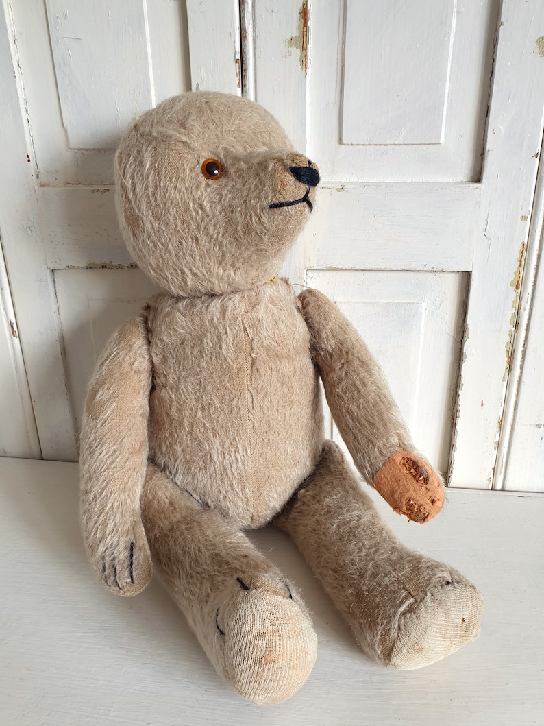 May include: A vintage beige teddy bear with black eyes and a stitched nose. The bear is sitting with its legs crossed and has a worn patch on one arm.