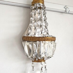 May include: An antique wall sconce with a gold-colored crown and trim, adorned with hanging crystal beads and rectangular crystal accents. The sconce is mounted on a white wall, offering a touch of vintage elegance.