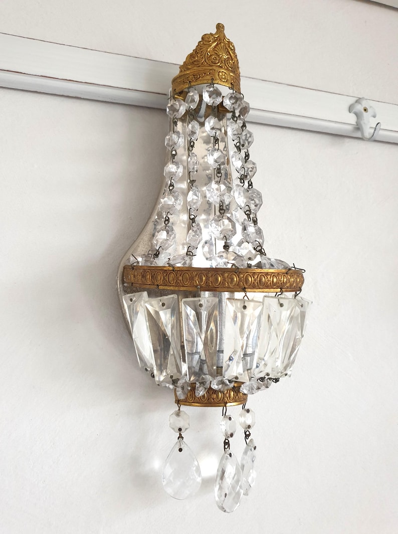 May include: An antique wall sconce with a gold-colored crown and trim, adorned with hanging crystal beads and rectangular crystal accents. The sconce is mounted on a white wall, offering a touch of vintage elegance.