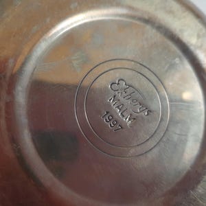 May include: Close-up of a metallic, possibly stainless steel, object. The center features a circular emblem with the text "Ekbergs MALM 1997" in a stylized font. The surface shows signs of wear.