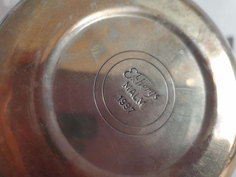 May include: Close-up of a metallic, possibly stainless steel, object. The center features a circular emblem with the text "Ekbergs MALM 1997" in a stylized font. The surface shows signs of wear.