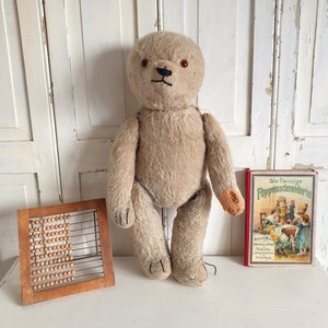 May include: A beige teddy bear with black eyes and a brown nose sits on a white surface. A wooden abacus and a red book with the title "Die fleissige Puppenscheiderin" are also in the image.