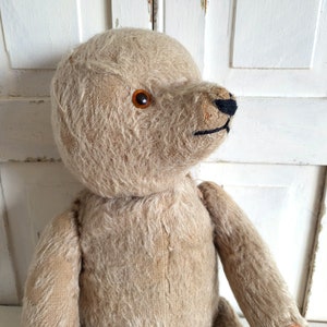 May include: A vintage, beige teddy bear with brown button eyes and a black stitched nose. The bear is made of soft, plush fabric and has a worn, well-loved appearance.