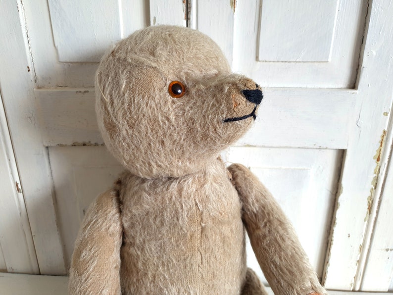 May include: A vintage, beige teddy bear with brown button eyes and a black stitched nose. The bear is made of soft, plush fabric and has a worn, well-loved appearance.