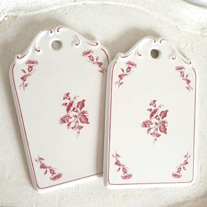 Könnte beinhalten: Zwei weiße Keramik-Schneidebretter mit rotem Blumendesign. Jedes Brett hat eine dekorative Oberseite mit einem Loch zum Aufhängen. Die Bretter haben einen roten Rand und florale Akzente.