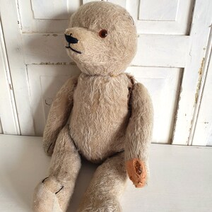 May include: A vintage, beige teddy bear with one brown eye and a black stitched nose. The bear is sitting with its legs outstretched and has some wear and tear on its fur and stitching.