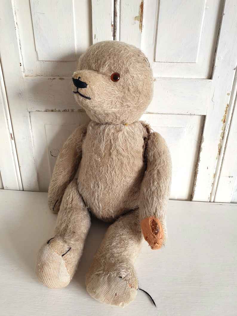 May include: A vintage, beige teddy bear with one brown eye and a black stitched nose. The bear is sitting with its legs outstretched and has some wear and tear on its fur and stitching.