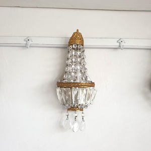 May include: Three antique crystal wall sconces with gold-colored metal accents. Each sconce features a crown-like top, a cascade of clear crystals, and hanging crystal pendants. The sconces are mounted on a white wall.