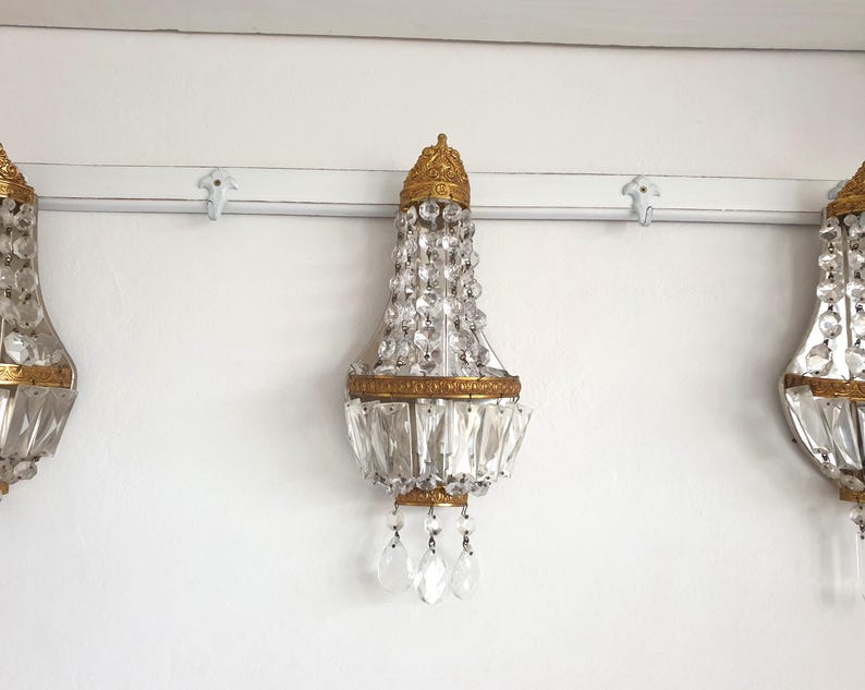 May include: Three antique crystal wall sconces with gold-colored metal accents. Each sconce features a crown-like top, a cascade of clear crystals, and hanging crystal pendants. The sconces are mounted on a white wall.