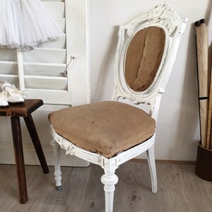 Puede incluir: Silla de madera pintada de blanco con un asiento de tela marr&oacute;n y tallas ornamentadas. La silla tiene un respaldo alto y un asiento curvo.