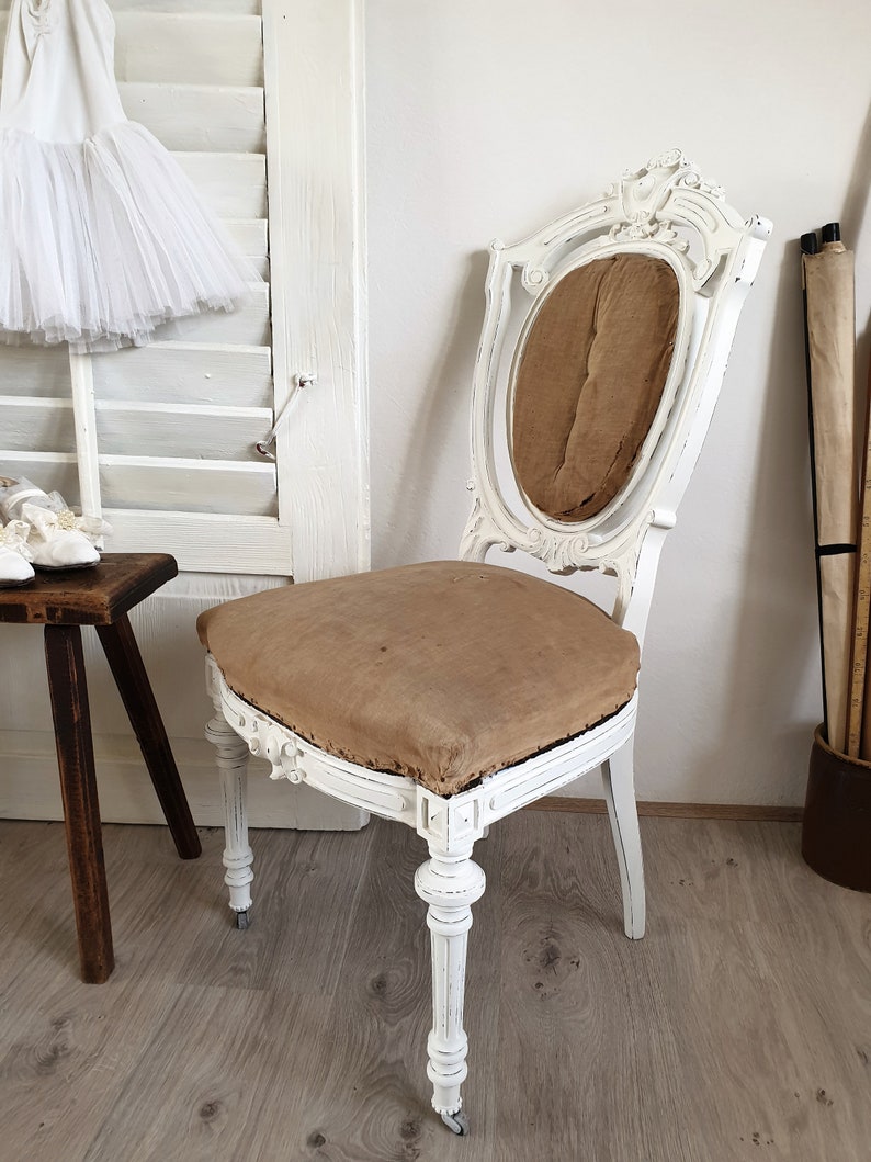 Puede incluir: Silla de madera pintada de blanco con un asiento de tela marr&oacute;n y tallas ornamentadas. La silla tiene un respaldo alto y un asiento curvo.