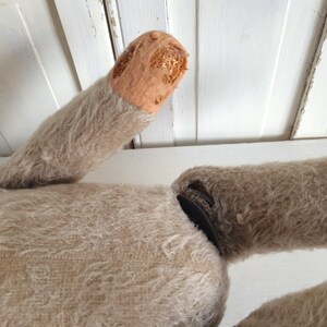May include: A vintage, beige, furry toy with a missing arm. The arm is visible, showing the stuffing inside. The toy is lying on a white surface.