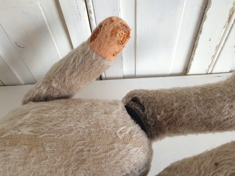 May include: A vintage, beige, furry toy with a missing arm. The arm is visible, showing the stuffing inside. The toy is lying on a white surface.