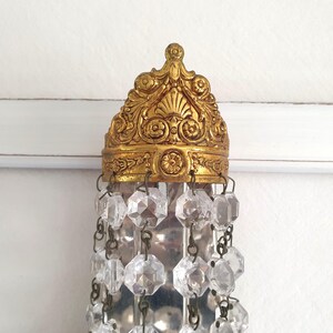 May include: Ornate gold-colored chandelier fixture with clear crystal accents. The fixture has a detailed, decorative crown-like top and hanging crystal strands. The crystals are faceted and reflect light, adding sparkle to the design.