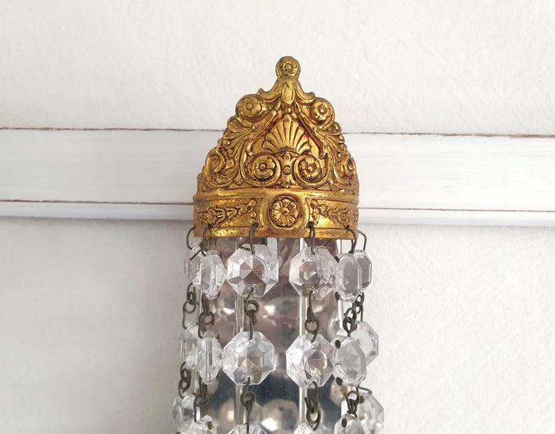 May include: Ornate gold-colored chandelier fixture with clear crystal accents. The fixture has a detailed, decorative crown-like top and hanging crystal strands. The crystals are faceted and reflect light, adding sparkle to the design.