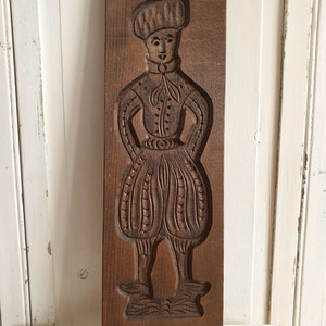 Puede incluir: Un molde de galletas de madera vintage con una figura tallada de una persona con vestimenta hist&oacute;rica. El molde marr&oacute;n oscuro representa a una persona con sombrero, chaqueta y pantalones abombados. El molde es rectangular y parece bien conservado.