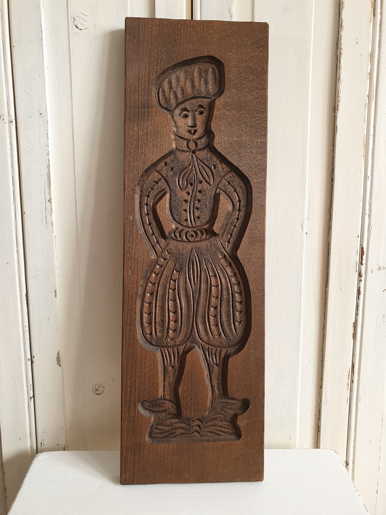 Puede incluir: Un molde de galletas de madera vintage con una figura tallada de una persona con vestimenta hist&oacute;rica. El molde marr&oacute;n oscuro representa a una persona con sombrero, chaqueta y pantalones abombados. El molde es rectangular y parece bien conservado.