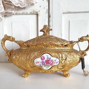 May include: An antique gold-coloured decorative container with a lid, handles, and feet. The container has a floral design on a white oval insert. The handles are dragon-shaped. A black tag hangs from the handle.