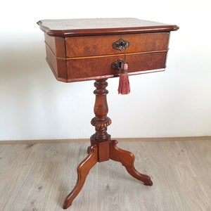 Könnte beinhalten: Ein antiker Beistelltisch aus Holz mit zwei Schubladen und einem einzelnen Sockel. Der Tisch hat eine dunkelbraune Farbe und aufwendige Schnitzereien an den Beinen. Ein rosa Quaste hängt an der unteren Schublade.