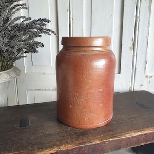 May include: A vintage, cylindrical, brown stoneware jar with a lid. The jar has a slightly glossy finish and embossed text on the front. It sits on a rustic wooden stool. A vase of dried lavender is in the background.