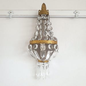 Puede incluir: Aplique de pared antiguo con marco de metal dorado y colgantes de cristal en cascada. El aplique presenta una parte superior e inferior decorativas, con m&uacute;ltiples elementos de cristal transparente. Montado en una pared blanca.