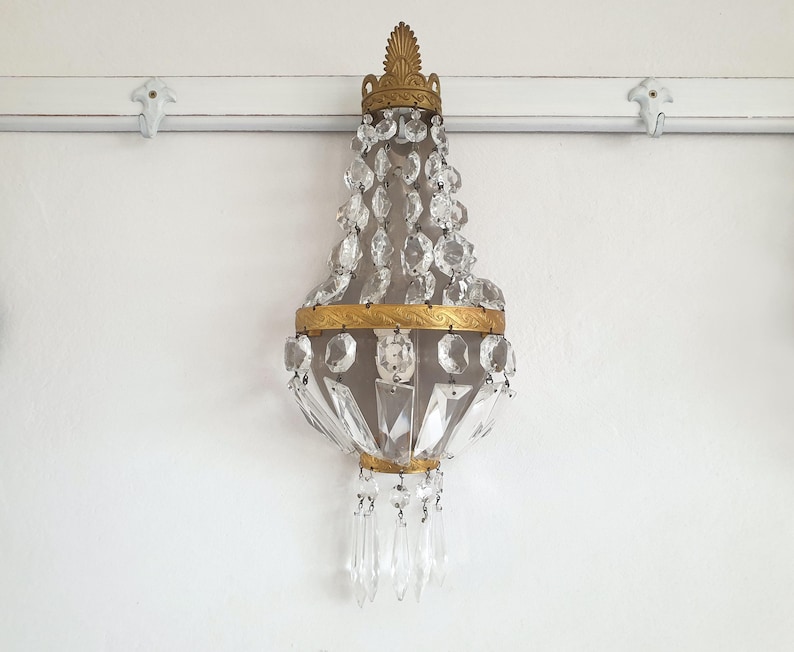 Puede incluir: Aplique de pared antiguo con marco de metal dorado y colgantes de cristal en cascada. El aplique presenta una parte superior e inferior decorativas, con m&uacute;ltiples elementos de cristal transparente. Montado en una pared blanca.