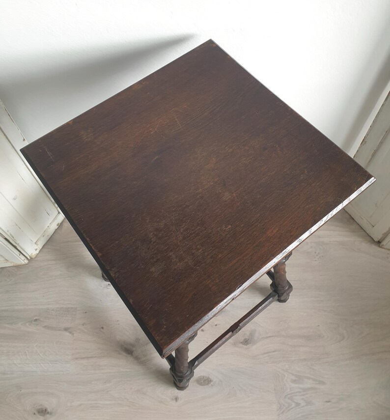 May include: A dark brown, square side table with a distressed finish. The table has four ornate legs and a connecting stretcher. The tabletop is approximately 24 inches square.