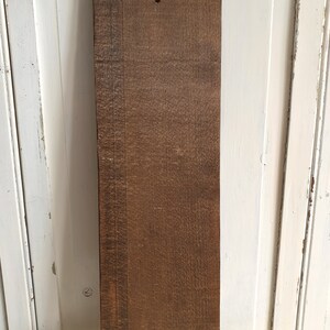 Puede incluir: Una tabla de madera rectangular con un color marr&oacute;n intenso y veta visible. La tabla tiene un agujero perforado cerca de la parte superior y mide aproximadamente 76 cm de largo. La madera parece envejecida y desgastada.