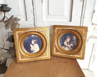 May include: Two antique framed portraits with gold-colored frames. The round portraits depict children, one praying and the other holding a dog. The frames have ornate detailing and are set on a wooden surface.