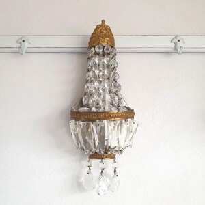 May include: An antique wall sconce with a gold-colored metal frame and clear crystal accents. The sconce has a decorative top and bottom, with hanging crystal pendants. The fixture is mounted on a white wall.