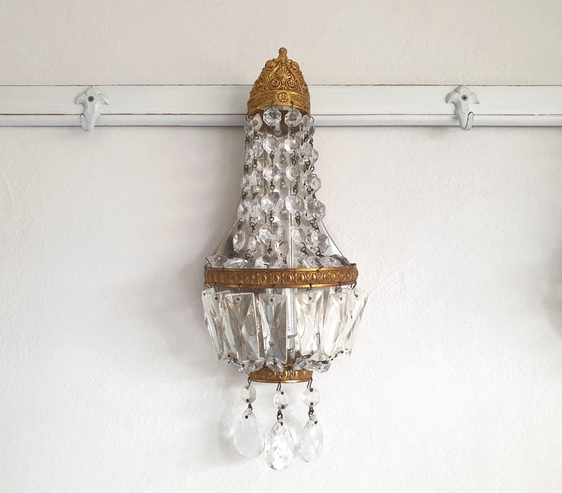 May include: An antique wall sconce with a gold-colored metal frame and clear crystal accents. The sconce has a decorative top and bottom, with hanging crystal pendants. The fixture is mounted on a white wall.